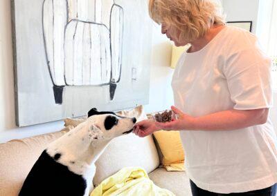 Sonya Weisshappel stands in front of the couch in her living room, holding a bowl of dog treats. She is feeding one treat to her dog, Finn