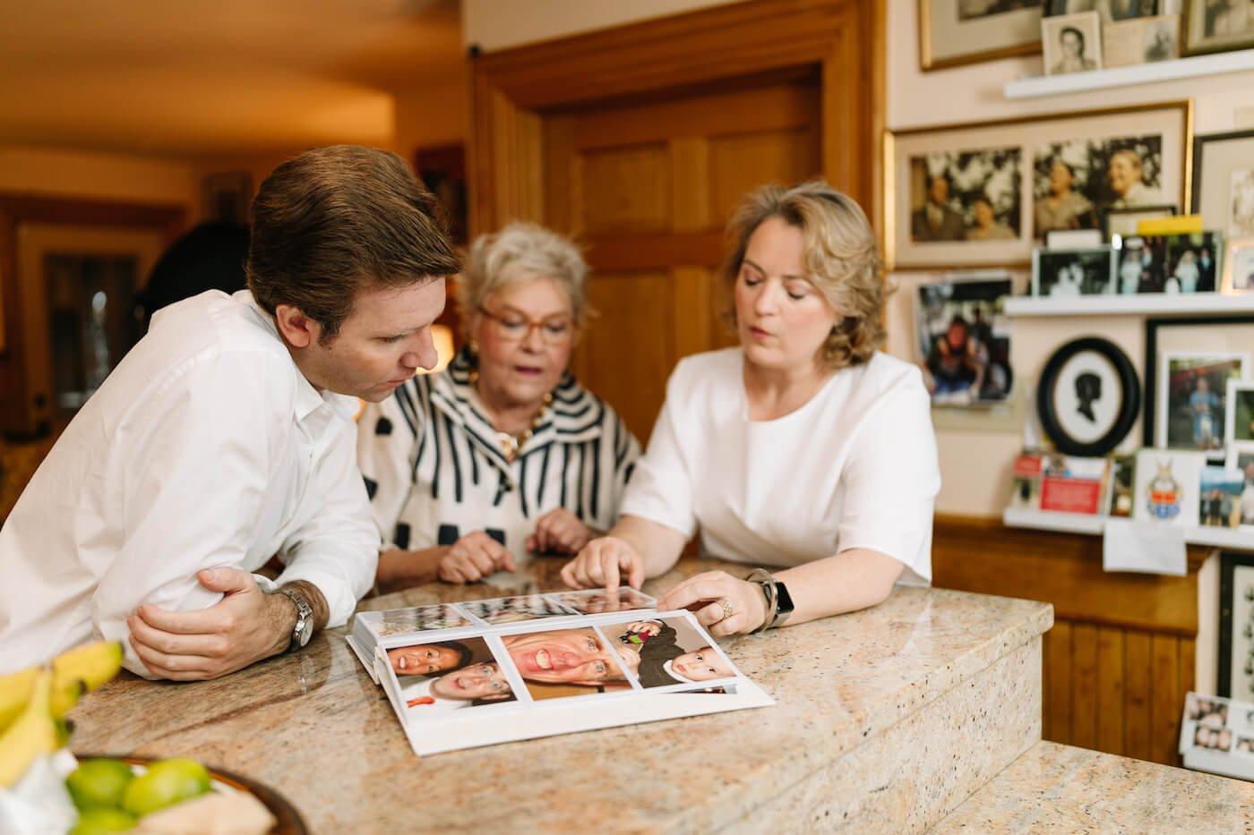 Haas Regen, Sonya Weisshappel, and a Seriatim client sit at a kitchen counter with a family photo album open in front of them. They are discussing the contents of the album.