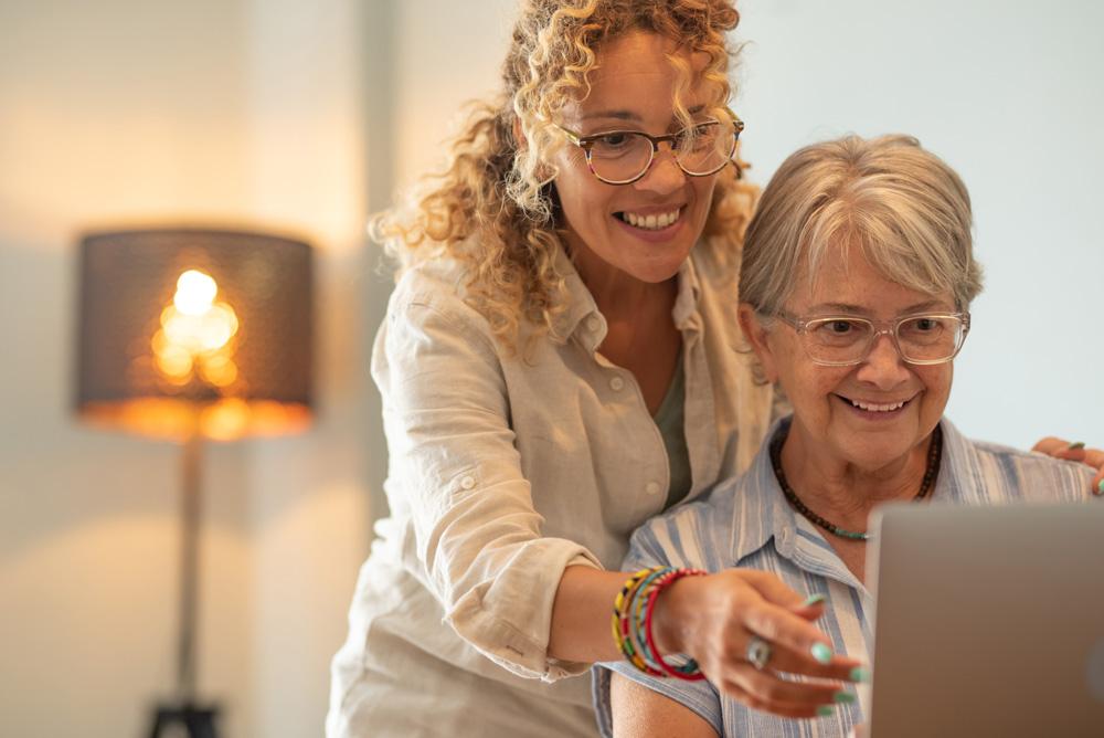 A professional moving coordinator helps an older woman use a laptop in a well-lit room, both smiling as they look at the screen.