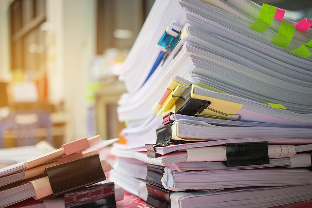 Stacks of documents and reports held together with binder clips and sticky notes on a cluttered office desk.