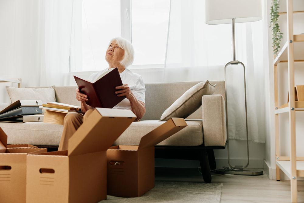 A elderly woman sits on a couch in a well-lit living room. She holds a photo album in her hands and cardboard boxes crowd the floor at her feet