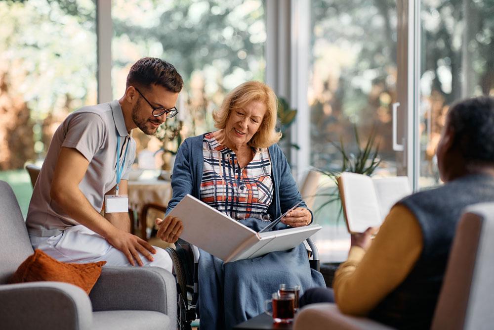 A man and an elderly woman in a wheelchair look at a photo album together, while another person reads a book nearby in a bright, home-like assisted-living setting.