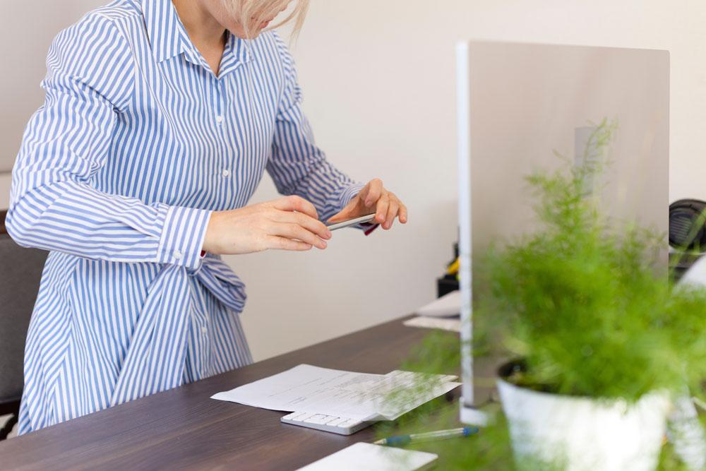 A person in a blue striped shirt stands in front of a desk, digitizing a document by taking a photo with a smartphone.