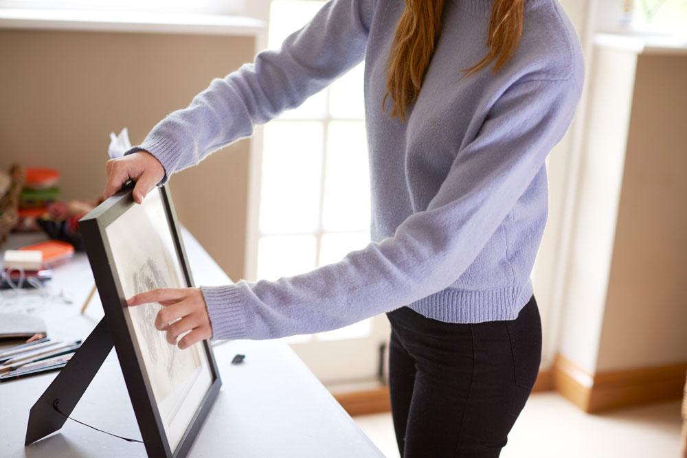 A person in a light purple sweater adjusts a framed picture on a table in a sunlit room.