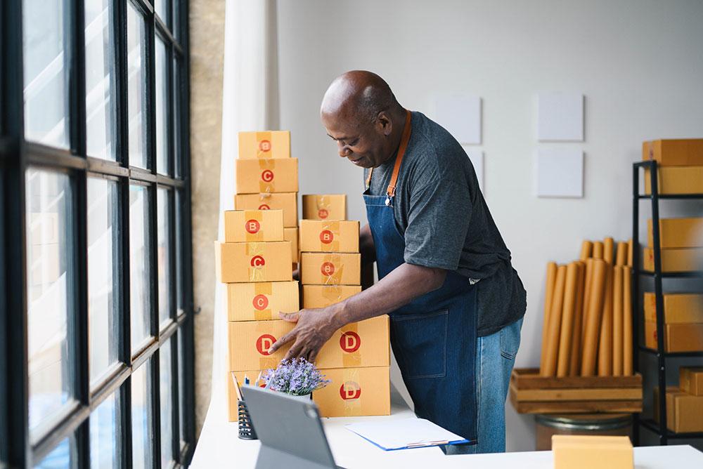 A man in an apron arranges stacked cardboard boxes on a table in a workspace with packing materials and a laptop nearby.