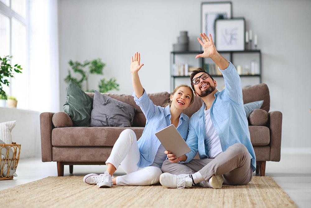 Two people sit on a rug in a living room, smiling and looking upward as though envisioning moving into their new home, with a tablet in hand. A sofa and decor are visible in the background.