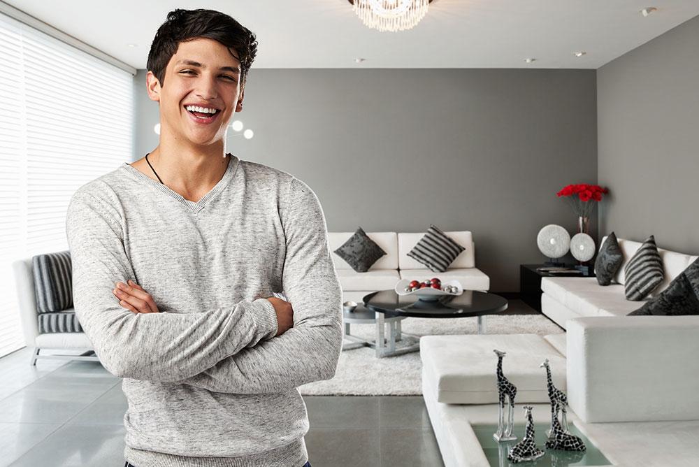 Young man with dark hair and a gray long-sleeve shirt stands smiling with arms crossed in a modern, stylish living room with light-colored furniture and decorative accents.