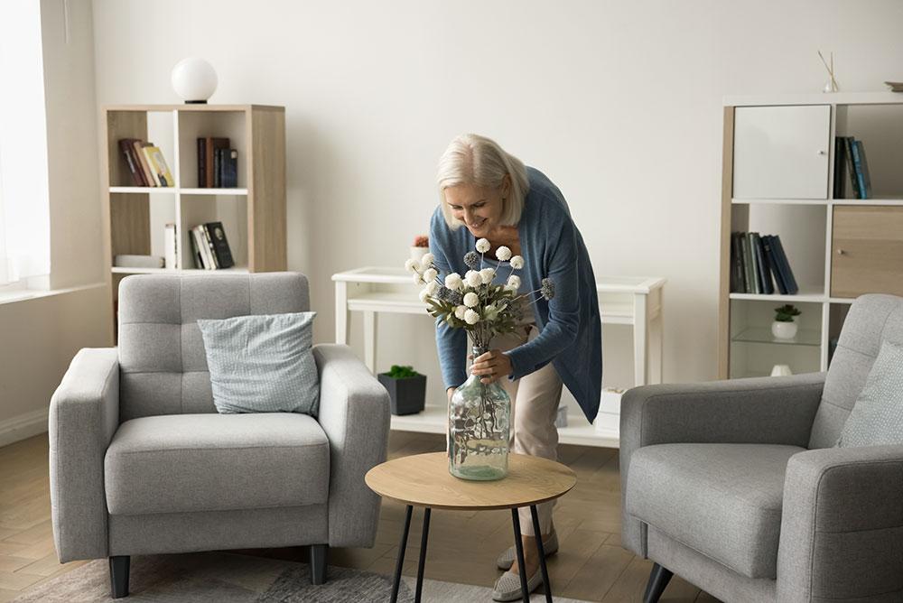 A professional organizer carefully places a vase with white flowers on a small round table between two gray armchairs in a bright, modern living room.