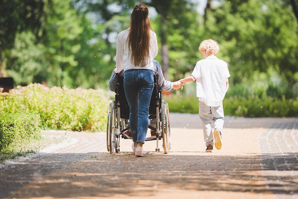 A woman pushes a person in a wheelchair along a park path while a young child walks beside them, holding the wheelchair users hand.