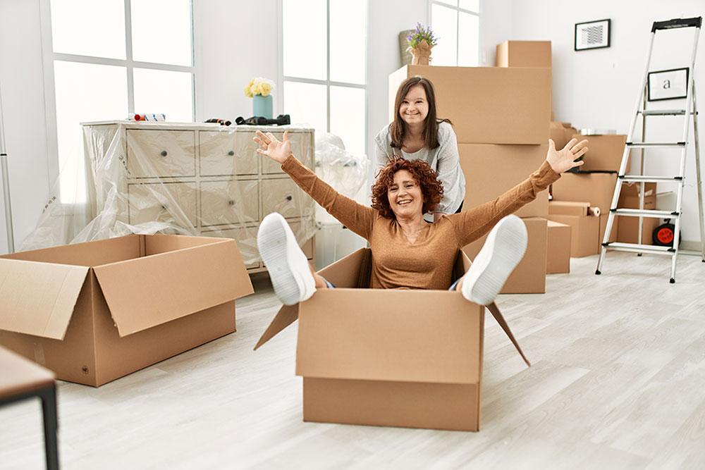 Two women in a room with moving boxes; one sits inside a box with arms raised, while the other stands behind, both appearing happy.