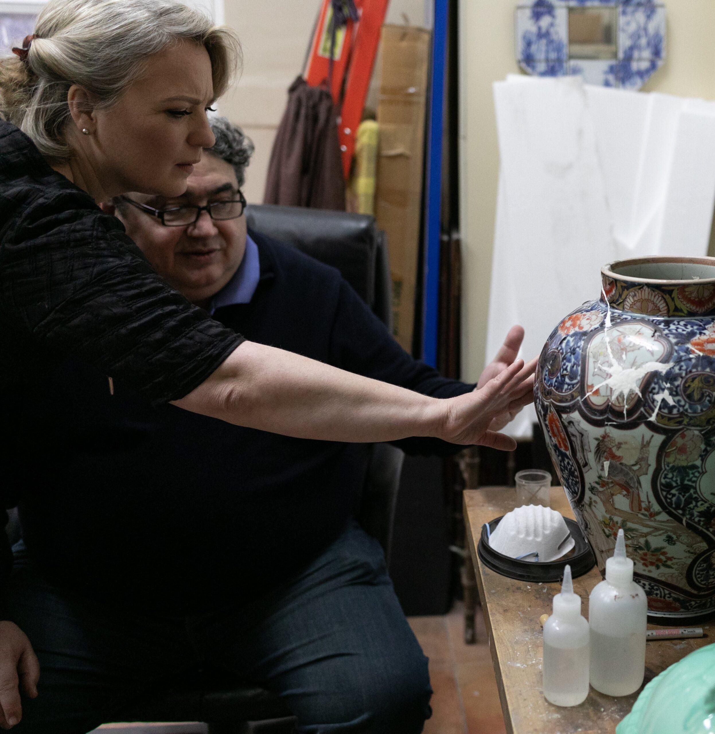 2W8A2646 Sonya Weisshappel examines a large, ornate ceramic vase while a man sits behind her, explaining his work. The vase is on a bench in a restoration workshop; beside it sit various supplies and a dust mask.