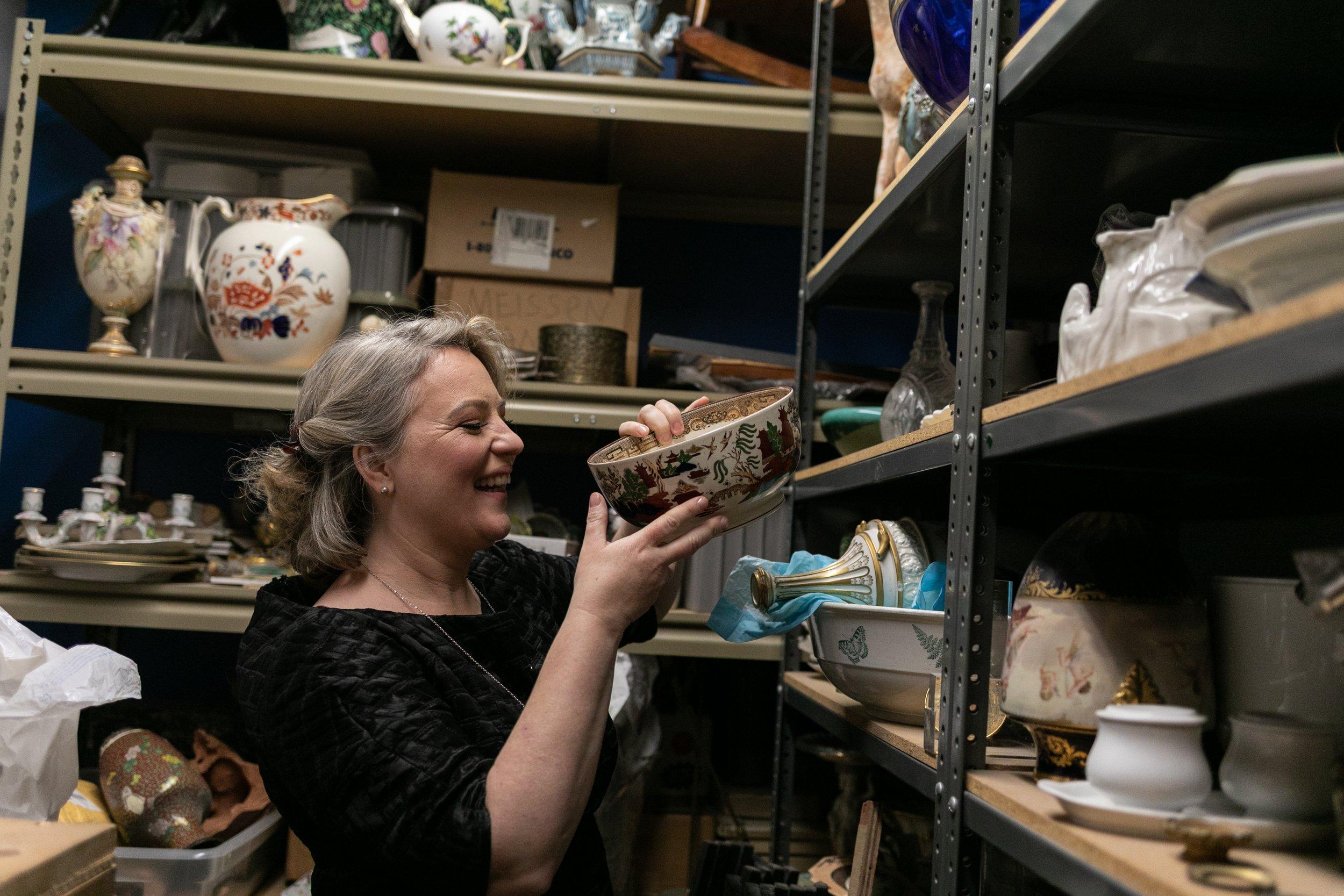 Sonya Weisshappel, professional organizer and transition support expert, stands in a storage room filled with shelves and various family heirlooms. She holds a china bowl and smiles joyfully.