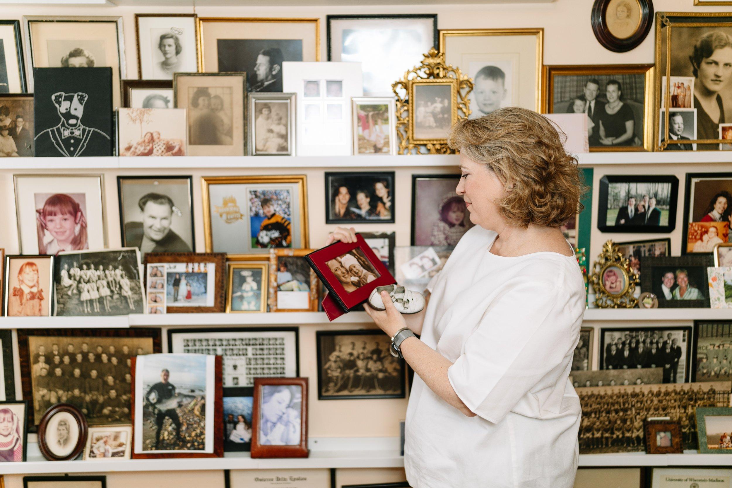 Sonya Weisshappel stands in front of a wall of family photographs. She holds a photo in a frame and looks at it, smiling.
