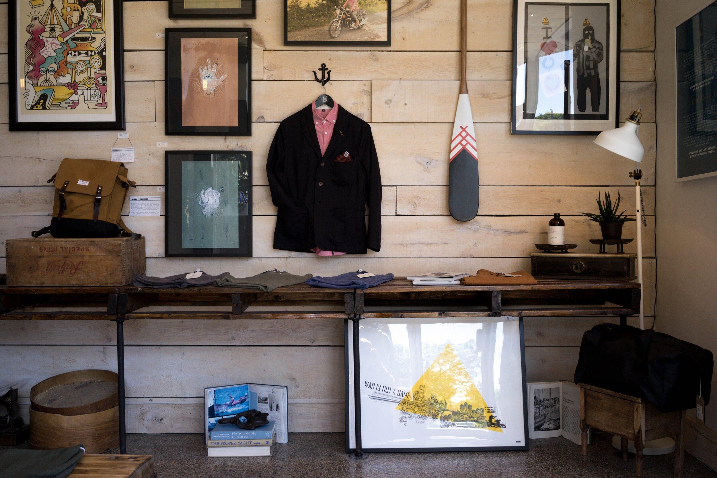 A black blazer and pink shirt hang on a wooden wall surrounded by framed artwork above a table displaying Artful Ephemera—clothes, bags, and decorative items—in a shop.