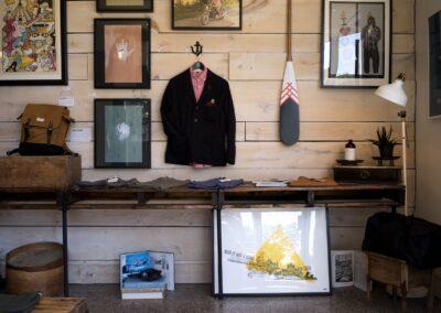 A black blazer and pink shirt hang on a wooden wall surrounded by framed artwork above a table displaying Artful Ephemera—clothes, bags, and decorative items—in a shop.