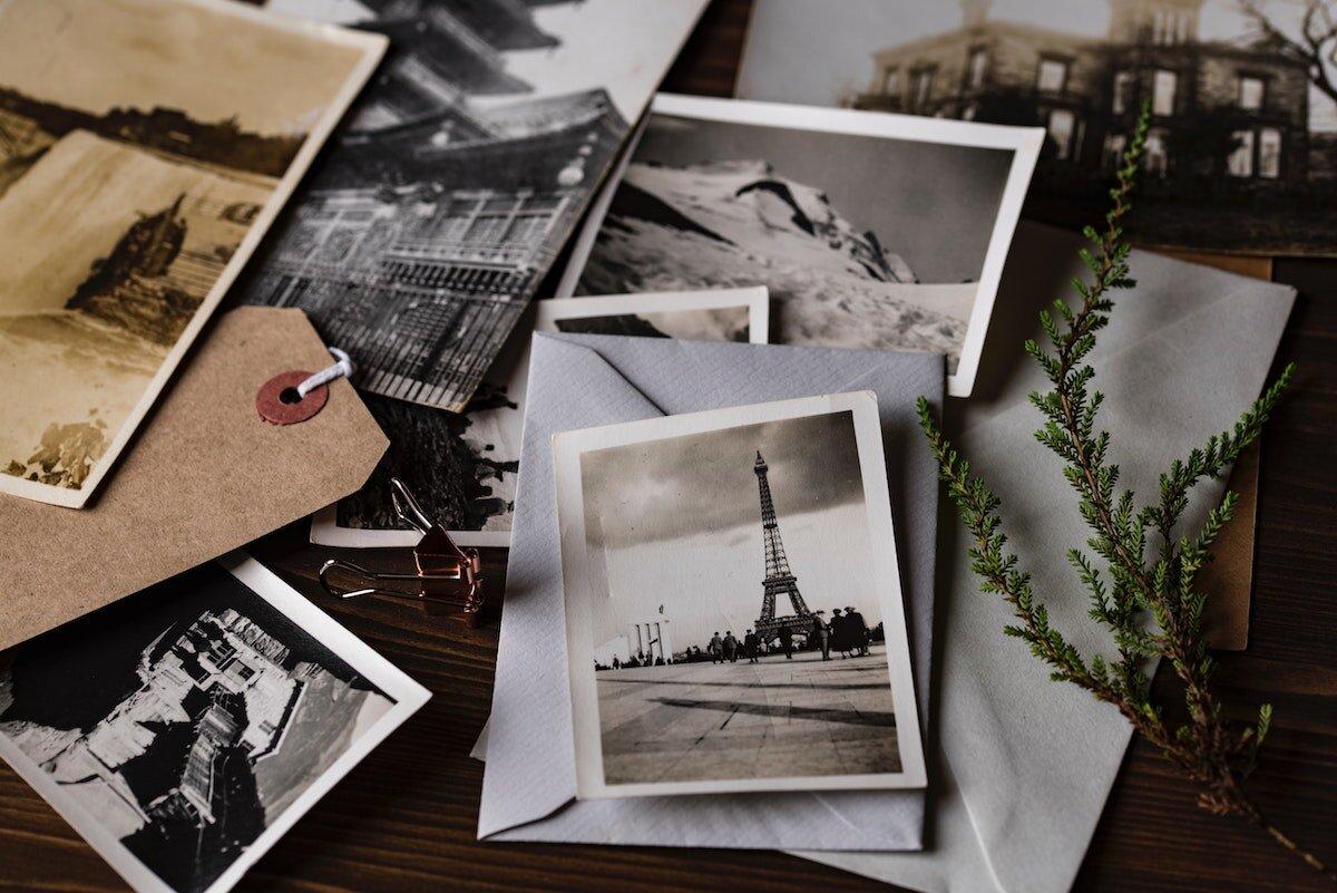A collection of vintage black and white photographs, perfect for photo archiving, including one of the Eiffel Tower, scattered on a table with envelopes, a sprig of greenery, and a brown tag.
