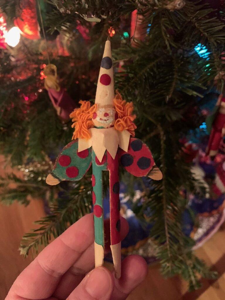 A hand holding a clown Christmas ornament made from a clothespin, painted with colorful polka dots and orange yarn hair, in front of a decorated Christmas tree.