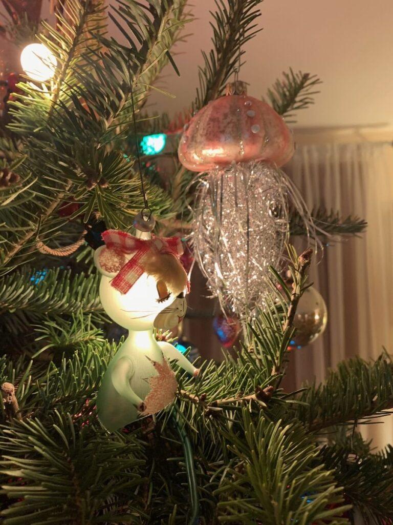 A close-up of a Christmas tree decorated with a cat ornament wearing a red hat and a shiny jellyfish ornament, with tree branches and lights in the background.