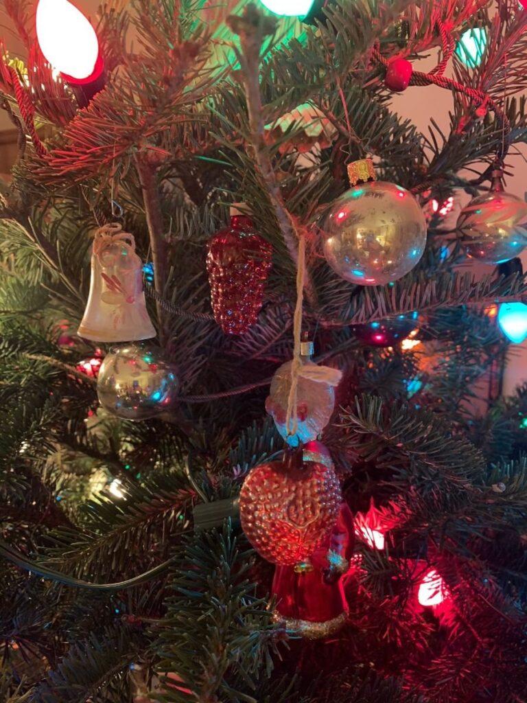 A close-up of a Christmas tree with colorful lights and various ornaments, including a bell, a Santa, a silver ball, and a red grape-shaped ornament.