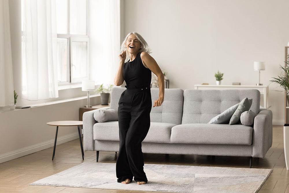 A woman with gray hair in a black outfit dances barefoot on a rug in a bright living room with a gray sofa and large windows.