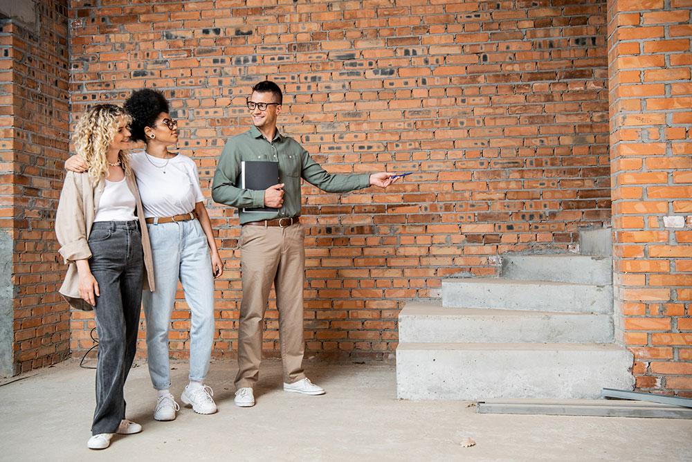 A man gestures toward a concrete staircase while two women stand together in front of an unfinished brick wall.