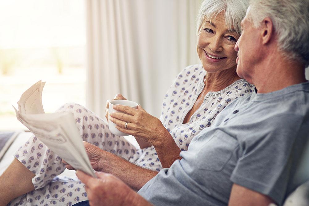 An older woman holding a mug smiles at an older man who is reading a newspaper; they are sitting together indoors in casual clothes.