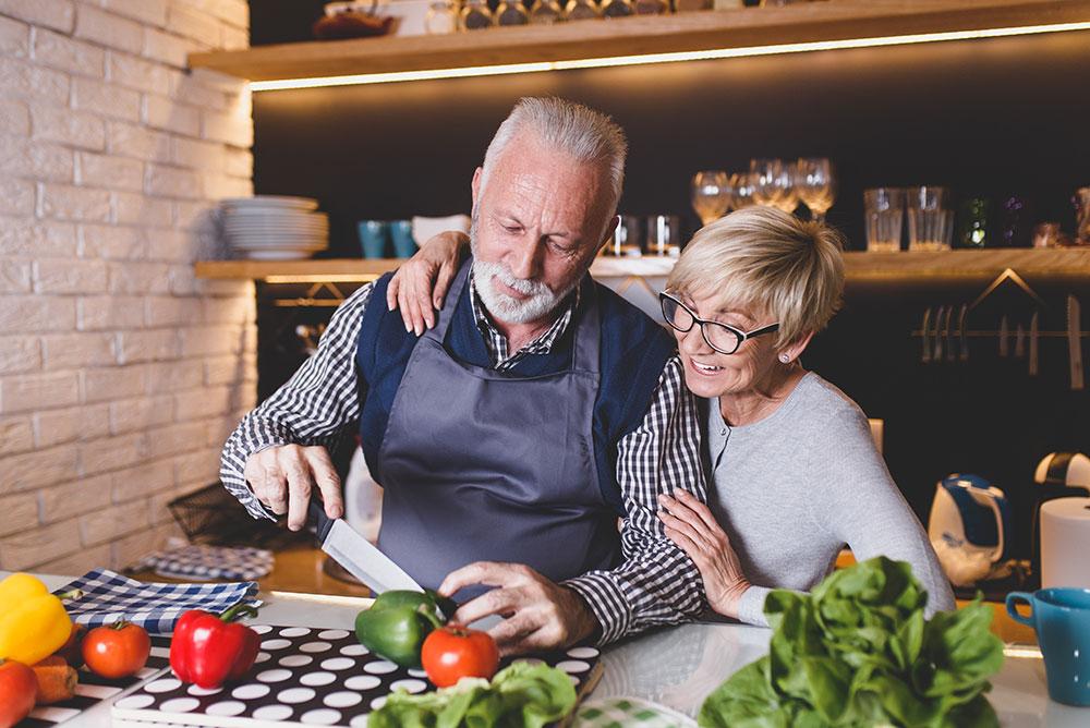 An older man slices a green bell pepper on a cutting board while a smiling woman stands beside him in a modern kitchen.