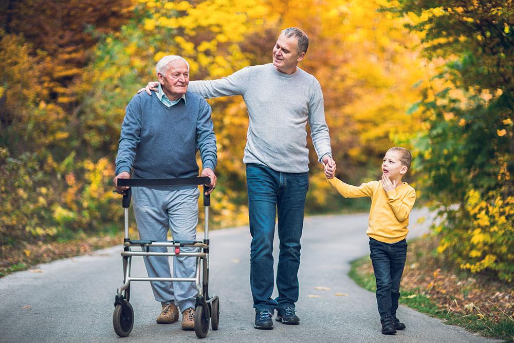 An elderly man with a walker, a middle-aged man, and a young boy walk together on a paved path surrounded by autumn trees.