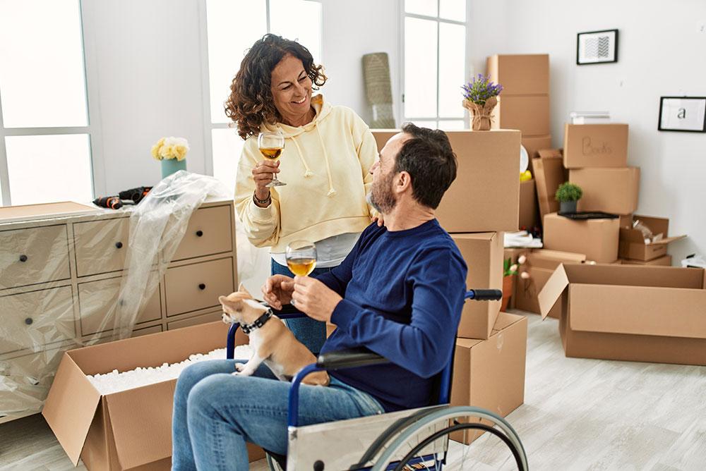 A woman and a man in a wheelchair, holding drinks and smiling at each other, sit among moving boxes in a bright room with a small dog on the man’s lap.