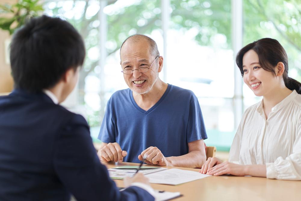 An older man and a younger woman sit together at a table, smiling and talking with a wealth manager in a suit, with documents in front of them. Bright windows and greenery are visible in the background.