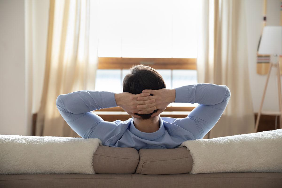 Person sitting on a couch with hands behind head, facing a window with light curtains and natural daylight entering the room.